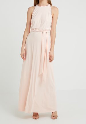 Kvinde iført en ærmeløs, lyserød maxikjole med taljebånd og åbne nudefarvede hæle, stående mod en ensfarvet lys baggrund.