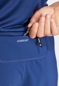 Modré športové šortky s celozipsovým vreckom, s logom "AEROREADY" a čiernou látkovou etikou. Hladká textúra a priliehavý strih.