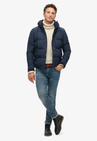 Chaqueta de plumas azul marino con capucha y cierre de botón, sobre un suéter de cuello alto color crema, combinada con jeans azules y botas negras con cordones.