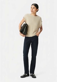 Beige kurzärmliges Strickoberteil, dunkelblaue eng anliegende Hose, hält eine schwarze strukturierte Handtasche. Schwarze Schuhe vervollständigen das Outfit.