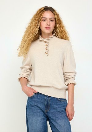 Ung kvinna med långt lockigt blont hår iklädd en beige volangtröja med knappar och blå jeans, stående med ena handen i fickan.
