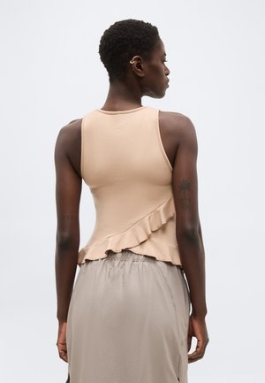 Moteris su trumpais garbanotais plaukais, vilkinti rankovių neturinčią smėlio spalvos palaidinę su raukiniais ir derančią lengvos pilkos spalvos sijoną, matoma iš nugaros.