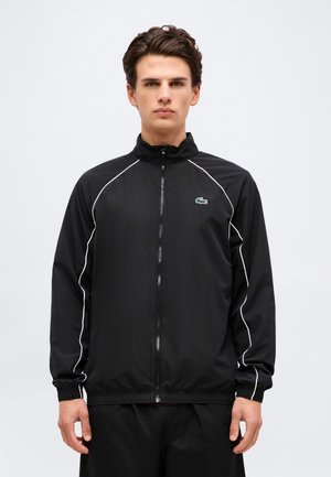 Giovane uomo con giacca nera con zip, piping bianco e piccolo logo di coccodrillo verde sul petto, in piedi davanti a uno sfondo uniforme.