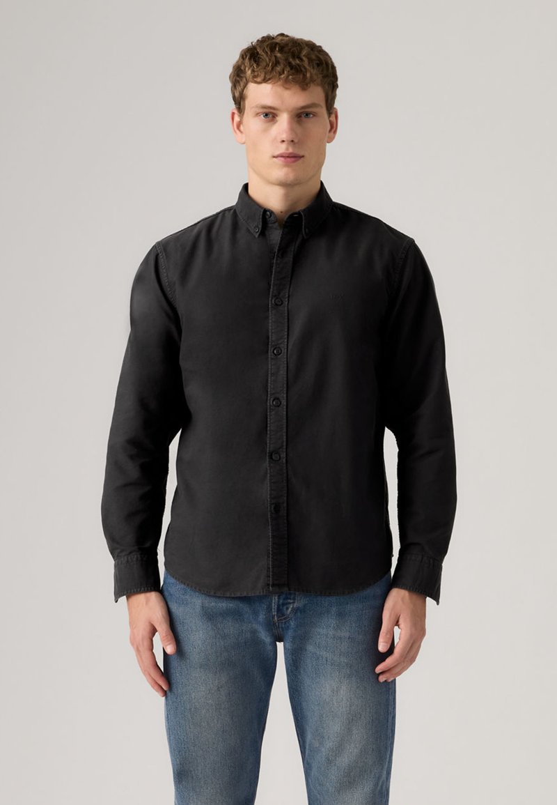 Μαύρο πουκάμισο με κουμπιά και μακριά μανίκια, κατασκευασμένο από απαλή ύφανση. Διαθέτει γιακά και διακριτική ραφή, σε συνδυασμό με ανοιχτόχρωμο τζιν παντελόνι.