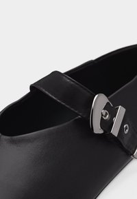 Chaussure Mary Jane en cuir noir avec boucle argentée sur le dessus, présentée en gros plan sur un fond clair.