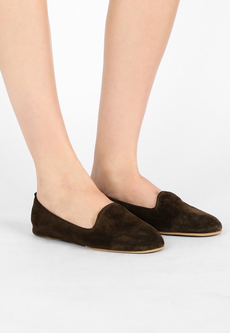 Pieds portant des chaussures plates en daim marron foncé avec un bout pointu arrondi et une fine semelle beige sur fond blanc.