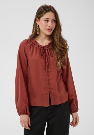 Femme aux longs cheveux ondulés porte un chemisier boutonné couleur rouille avec un col à nouer et des boucles d'oreilles anneaux dorés, associé à un pantalon noir, sur fond gris.