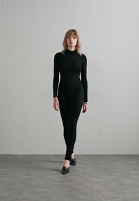 Black bodysuit with long sleeves, fitted design, smooth texture, paired with black flats. Model stands against a neutral background.