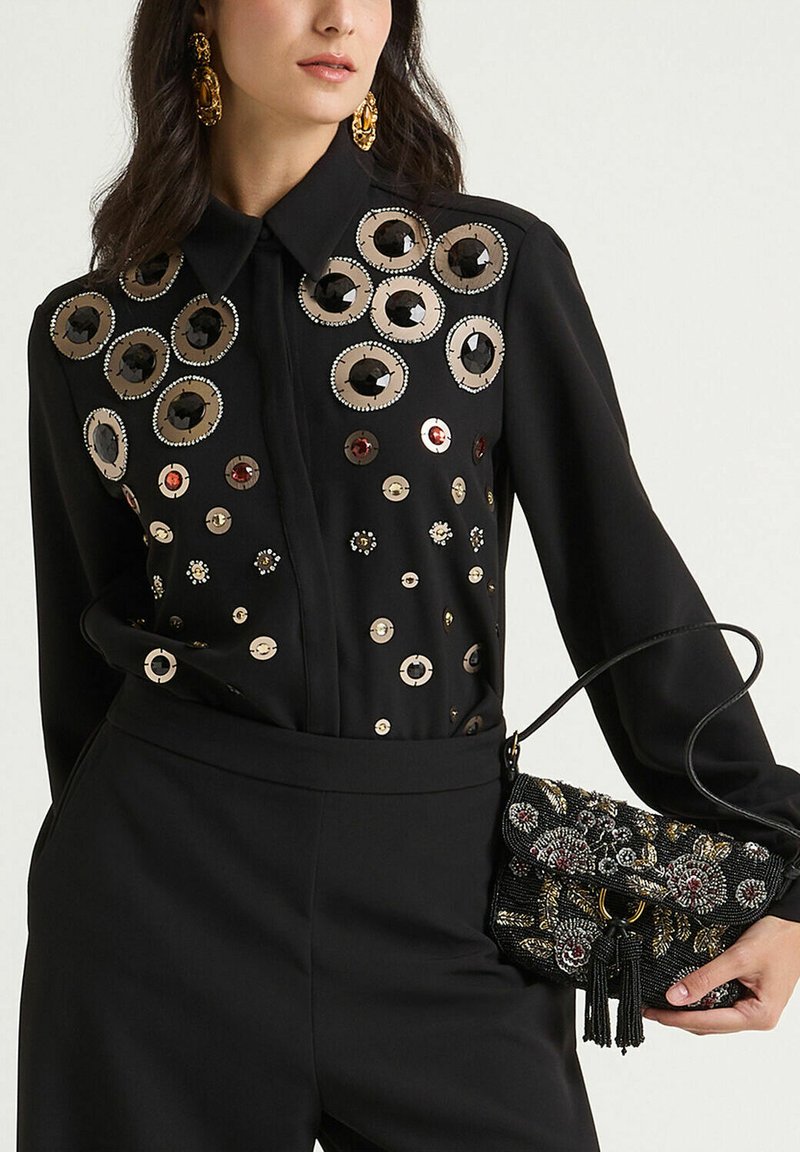 Blusa nera con decorazioni circolari metalliche e paillettes, abbinata a una piccola borsa nera con perline con motivi floreali e nappina.