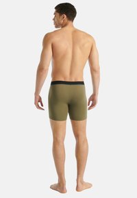 Muž stojící bos, otočený zády, v olivově zelených boxerkách s černým pasem na jednobarevném bílém pozadí.