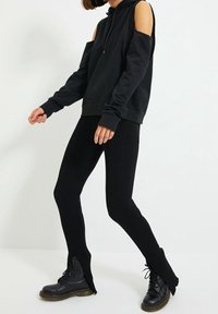 Zwarte hoodie met uitgesneden schouders, gecombineerd met geribde zwarte leggings. Beide kledingstukken hebben een aansluitende pasvorm, aangevuld met zwarte veterschoenen.
