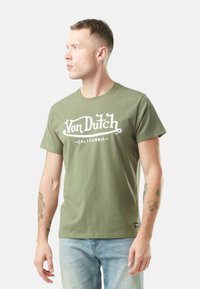 Homme avec les cheveux courts et des tatouages, portant un t-shirt vert Von Dutch California et un jean bleu clair, regardant vers la gauche sur un fond blanc.