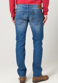 Blå denimjeans med rak ben, med en brun läderetikett på midjan och diskret blekta detaljer. Bakfickorna har rundad söm.