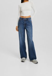 Wide-leg blue denim jeans with a high waist, featuring five pockets and a button closure. Paired with a white long-sleeve top.