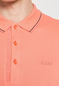 Gros plan d'une personne portant un polo rose saumon avec un col, trois boutons et « BOSS » brodé sur la poitrine.