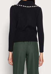 Maglione nero a maglia con colletto alto, orlo a costine e decorazioni floreali sul colletto. Indossato con pantaloni verdi.