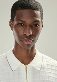 Gros plan d'un jeune homme aux cheveux courts portant un polo zippé blanc texturé, regardant directement l'appareil photo sur un fond uni.