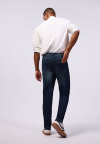 Jeans de mezclilla ajustados en azul oscuro con una textura suave, que cuentan con dos bolsillos traseros y una cinturilla, combinados con una camisa blanca de manga larga.