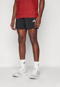 Tenue athlétique pour homme comprenant un t-shirt rouge rayé à séchage rapide, un short noir avec logo blanc, des chaussettes blanches et des baskets grises.