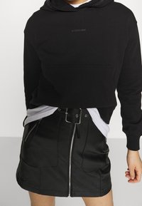 Sudadera negra corta con logo y bolsillo frontal, combinada con una falda mini de cuero negra con cremalleras y detalle de cinturón.