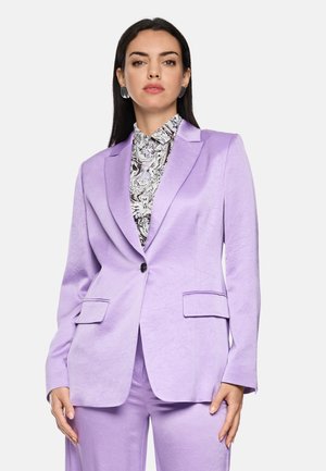 Woman wearing a light purple suit jacket and pants with a black and white patterned blouse, standing with a neutral expression.