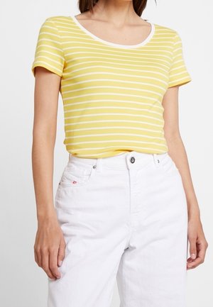 Kobieta w żółto-białej koszulce w paski z krótkim rękawem wpuszczonej w białe spodnie z wysokim stanem, stojąca z rękami luźno opuszczonymi.