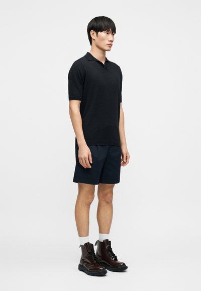 Polo en maille noire avec un col court, associé à un short bleu marine. Le mannequin porte des bottines à lacets marron et des chaussettes blanches aux chevilles.