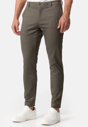 Smalbrune ternede bukser båret af en person parret med hvide lav-top sneakers, vist fra talje til fødder mod en ensfarvet baggrund.