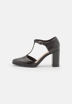 Chaussure haute à talon en cuir noir avec un bout en amande, design à bride en T et boucle réglable. Dispose d'un talon bloc solide.