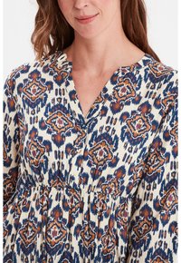 Gemusterte Bluse in Creme mit blauen, orangefarbenen und braunen Rautenmotiiven. Verfügt über einen V-Ausschnitt, Knopfakzente und eine geraffte Taille für eine schöne Form.