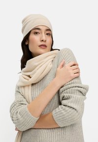 Beige gerippte Mütze und cremefarbener Schal, die über einem hellgrauen, strukturierten Pullover drapiert sind. Der Pullover hat lockere, aufgerollte Ärmel und eine lässige Passform.