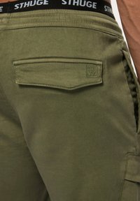 Shorts cargo vert olive en matériau doux, dotés d'une poche arrière avec rabat et du logo "STHUGE" en relief. Ceinture noire avec texte blanc.