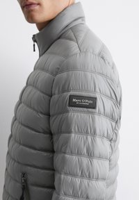 Grå vindtæt jakke med quiltet tekstur, høj krave og lynlås lomme. Har et mærke på ærmet med "Marc O'Polo" logo.