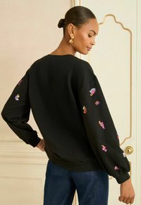 Melna sporta krekls ar apjomīgām piedurknēm, ar daudzkrāsainu fliteru ziedu akcentiem uz piedurknēm un noapaļotu kakla izgriezumu.