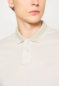 Beige poloing, texturált anyagból, két gombbal a gallérnál és fehér díszcsíkokkal. Diszkrét, kiemelkedő logó található a mellrészen.