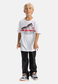 Blond chlapec stojí, má na sebe biely tričko s grafickým motívom, čierne nohavice a šedé tenisky s oranžovými akcentmi, vyplazuje jazyk hravo.