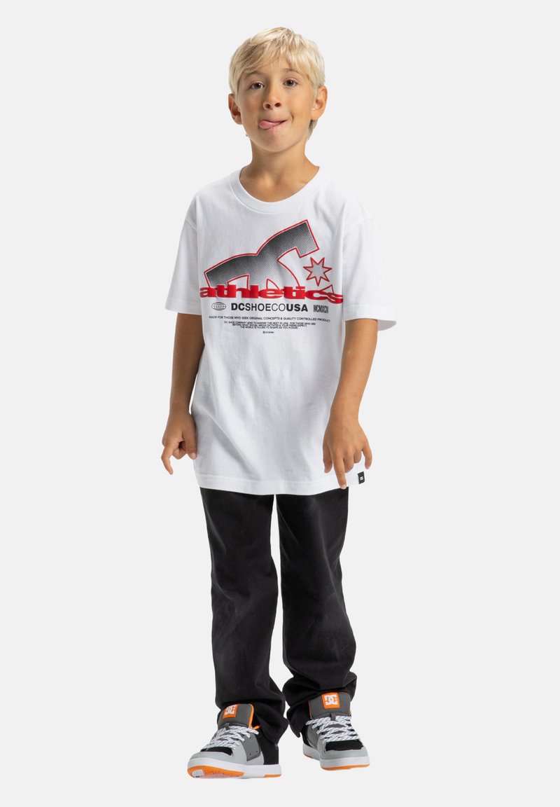 Blond chlapec stojí, má na sebe biely tričko s grafickým motívom, čierne nohavice a šedé tenisky s oranžovými akcentmi, vyplazuje jazyk hravo.