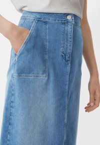 Osoba nosząca jasnoniebieską spódnicę dżinsową z dużą przednią kieszenią, ręka w kieszeni, a na wysokości talii widoczna biała bluzka.