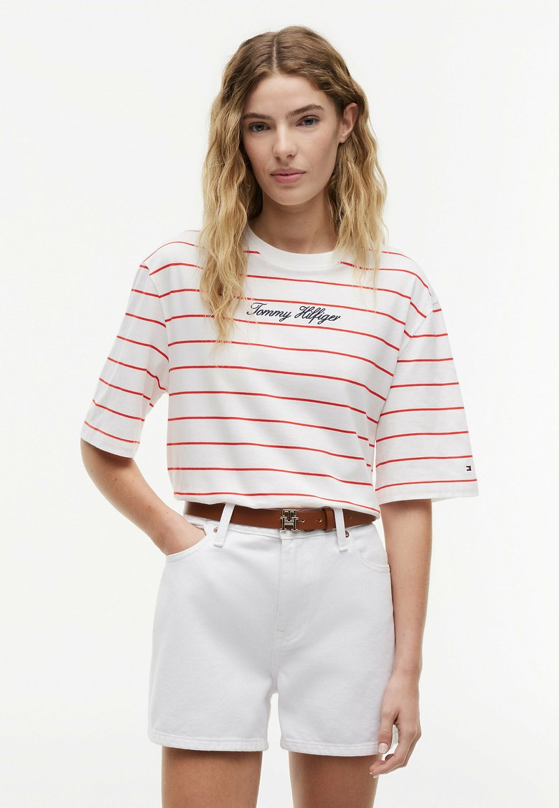 Femme aux cheveux blonds ondulés portant un t-shirt rayé blanc et rouge Tommy Hilfiger, un short blanc et une ceinture marron avec boucle "H".