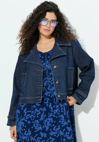 Riflová bunda tmavě modré barvy s krátkým střihem, velkým klopovým límcem, dvěma předními kapsami a kovovými knoflíky. Kombinována s modrými vzorovanými šaty.