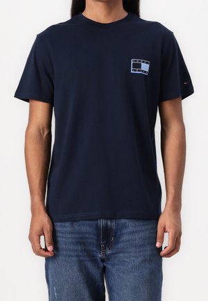 Mężczyzna w granatowej koszulce Tommy Jeans i niebieskich dżinsach, stojący na jednolitym białym tle.