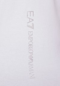 Bílé bavlněné tričko s vertikálním lesklým stříbrným textem "EA7 EMPORIO ARMANI" na boku, s měkkou texturou a standardní velikostí.