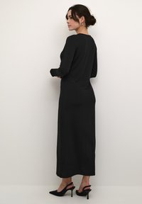Zwarte lange jurk met een getailleerde silhouette en subtiele schitterende versieringen. Draag met zwarte puntige hakken, getoond vanuit de achterkant.