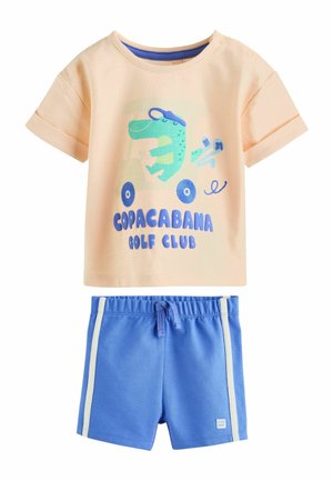 Bézs rövid ujjú kisgyermek póló kék dinóval egy golfkocsin és "Copacabana Golf Club" felirattal, kék rövidnadrággal fehér csíkokkal párosítva.