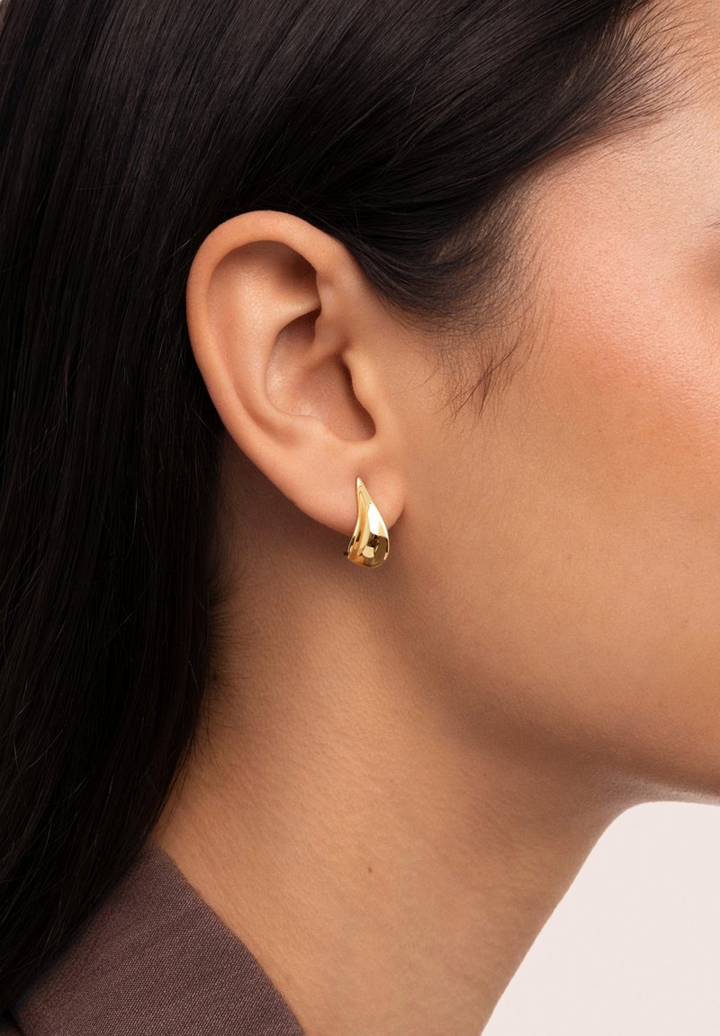 Goldene, tropfenförmige Ohrringe mit polierter Oberfläche, die am Ohr befestigt sind und glatte, reflektierende Flächen sowie ein elegantes Design präsentieren.