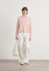 Pull en tricot rose pâle avec des poignets côtelés, associé à un large pantalon blanc. Le mannequin tient un sac fourre-tout crème ; textile lisse et coupe décontractée.