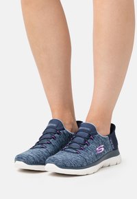 Scarpe sportive con una tomaia in tessuto blu navy, dettagli viola e lacci elastici. Suola leggera e collo imbottito per il comfort.