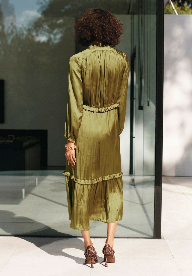 Femme aux cheveux bouclés portant une longue robe vert olive et des talons hauts imprimés léopard, debout devant une porte en verre.