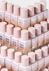 Et stortak med Revolution Skin Silk-flasker med firkantede baser, rosa lokk og kremfargede etiketter som har en gull-logo.