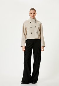 Beige gekürzte, doppelt knöpfbare Jacke mit schwarzen Knöpfen, weiten Ärmeln und strukturiertem Stoff, kombiniert mit schwarzen Cargo-Hosen mit seitlichen Taschen.